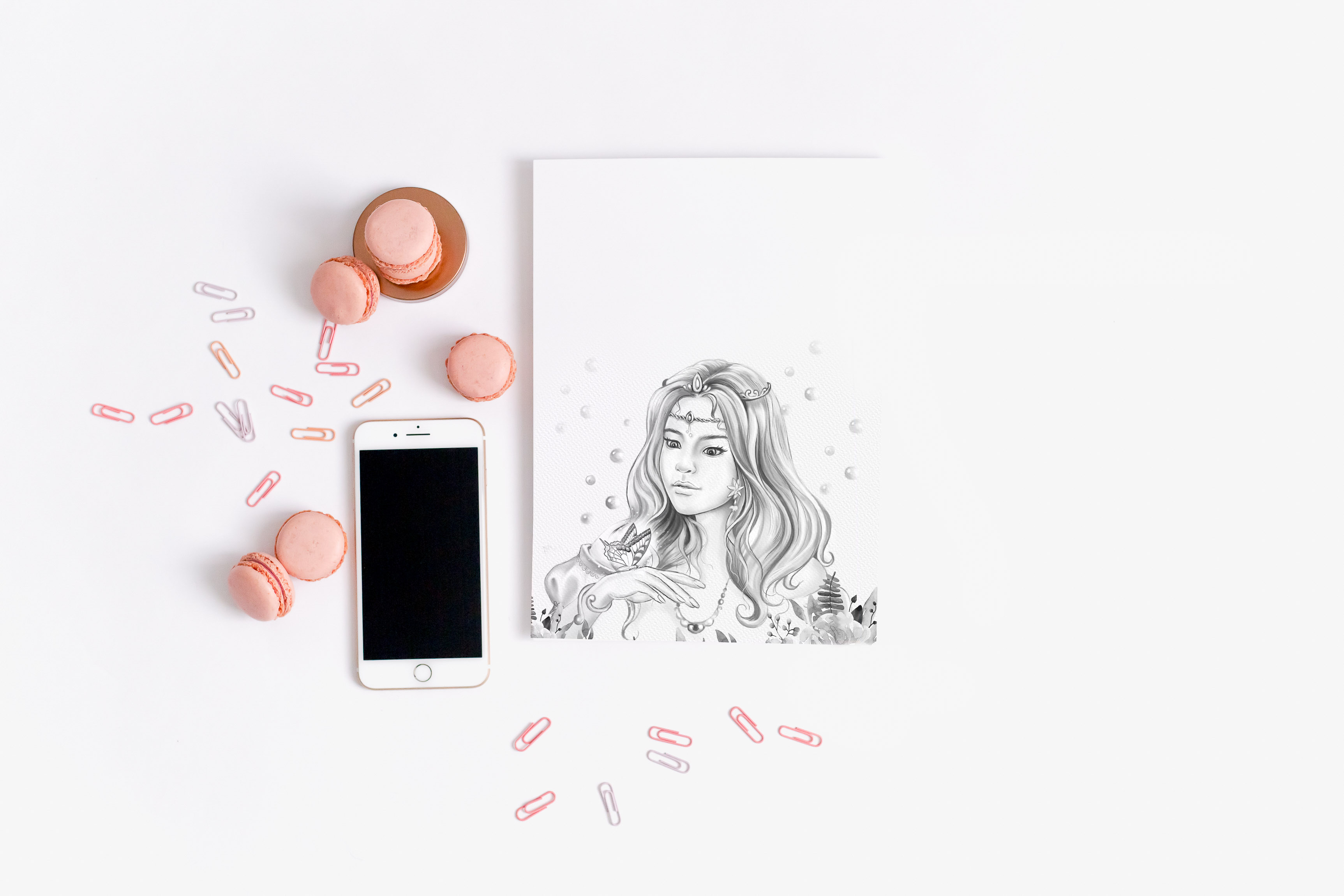 A coloring page of a young girl with long hair and a butterfly on her hand.  The page sits on a white background surrounded by a cell phone and pink macarons.